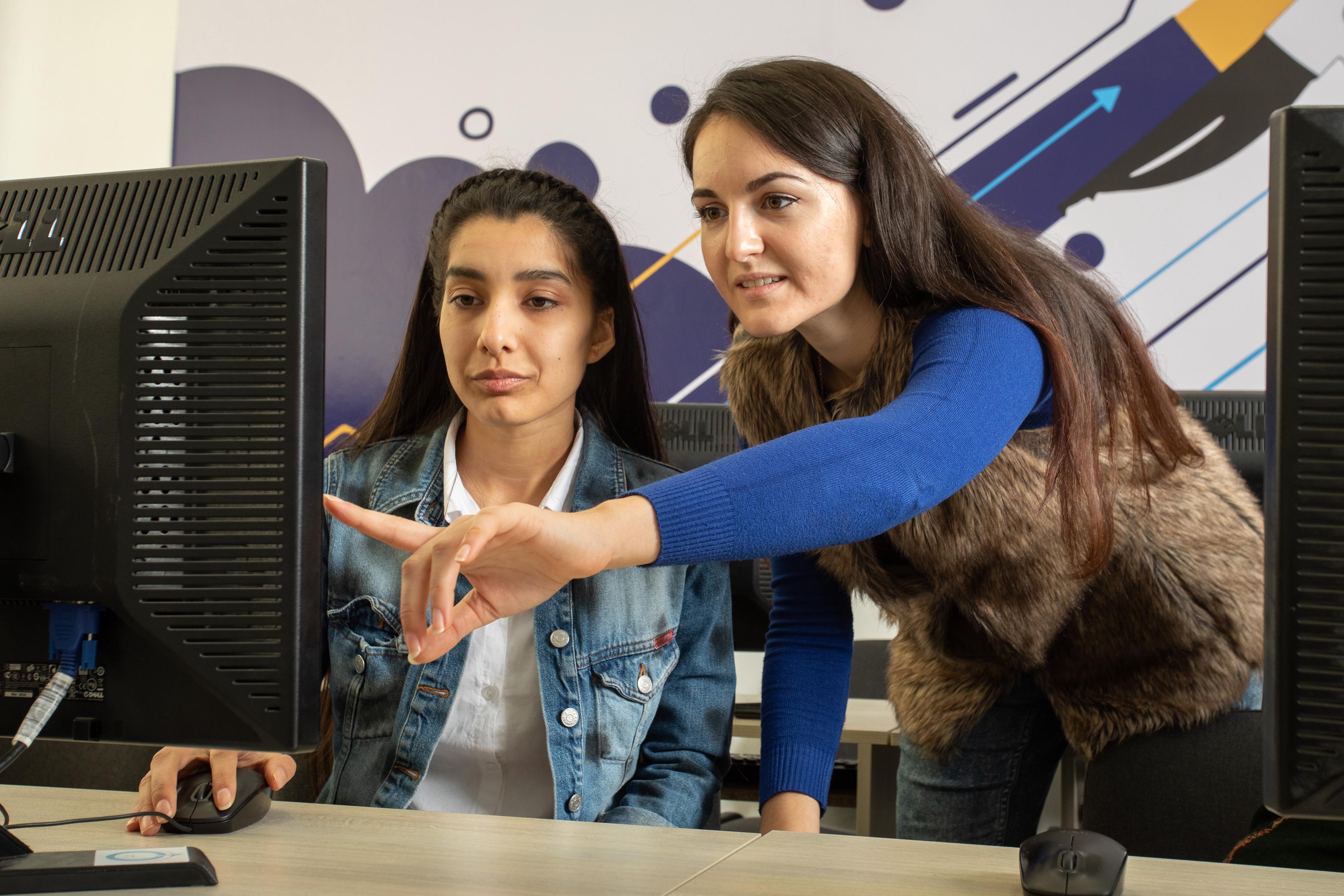 A young woman sitting in front of a computer in an office while another woman standing next to her is pointing at something on her screen. They are both dressed in casual clothes - a bright blue top and furry waistcoat, and a denim jacket.